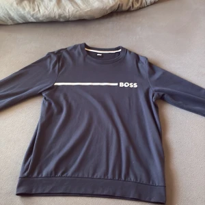 Svart långärmad tröja från BOSS - Svart långärmad tröja från BOSS med vit logga och linje framtill. Tröjan har rund halsringning och är tillverkad i mjuk bomull. Enkel och stilren design som passar perfekt till jeans eller chinos.