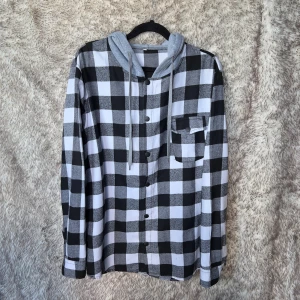 Svartvit rutig lumberjack skjorta med grå huva - Svartvit rutig lumberjack skjorta med grå huva och snörning. Skjortan har långa ärmar, knappar framtill, vid ärmsluten och en bröstficka med knapp. Materialet är mjukt och tunt.