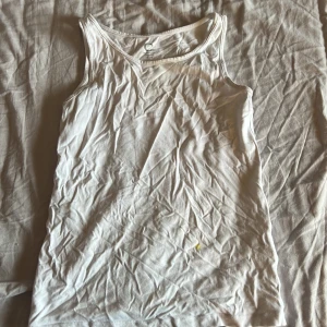 Vit ärmlös linne i bomull - En basic vit linnetopp från C i ekologisk bomull. Modellen är ärmlös med rund halsringning och har en enkel, rak passform. Polite skrynklig men bara att stryka! Liten gul fläck men syns inte när man har den på sig!