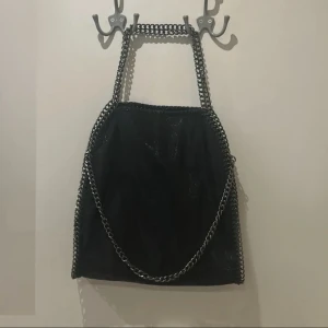 Svart axelväska med kedjedetaljer - Svart axelväska med silverfärgade kedjor längs kanterna och som axelrem. Väskan har en fyrkantig form. Nyskick har aldrig andvänt, säljer den för har två stycken
