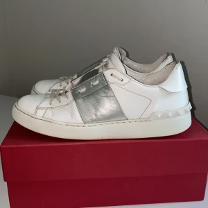 Valentino Opens  - Snygga vita Valentino Garavani Opens sneakers med metallic-silver detalj över vristen och ikoniska nitar på hälen. Skorna är i skinn och har snörning framtill. Perfekta för dig som vill ha en lyxig och trendig touch till din outfit.
