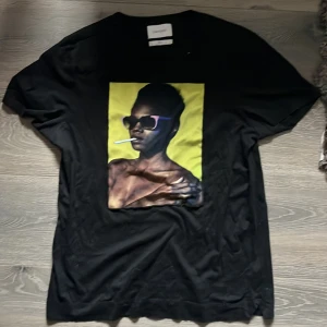 Svart Limitato t-shirt med konstmotiv - Svart t-shirt från Limitato med ett stort konstnärligt tryck på bröstet i gult och brunt. Klassisk passform och rund halsringning. Tillverkad i mjuk bomull och designad i samarbete med Riocam. Perfekt för dig som vill sticka ut med en unik look.