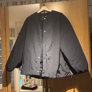 Svart oversized pufferjacka - Stilren svart pufferjacka med oversized passform och rund halsringning. Jackan har tryckknappar framtill och dragkedja i sidan för extra detalj. Perfekt för dig som gillar en enkel men trendig look. Aldrig använd 