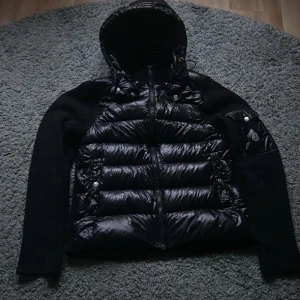 Svart pufferjacka från Moncler - Snygg svart pufferjacka från Moncler med glansig framsida och stickade ärmar. Jackan har huva, dragkedja framtill och en ficka med Moncler-logga på ärmen. Perfekt för dig som vill ha en stilren och exklusiv look under kyliga dagar.