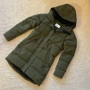 Grön jacka från Sail Racing - Snygg mörkgrön jacka från Sail Racing med huva och svarta detaljer. Jackan har två snedställda fickor med dragkedja och är quiltad för extra värme. Perfekt för kalla dagar och har en stilren, längre passform. Obs sminkfläckar på krage  förekommer men försvinner i tvätt. Se bilder