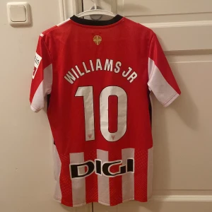 Athletic Bilbao matchtröja Williams Jr - Säljer en Athletic Club Bilbao fotbollströja från Castore, med Williams Jr och nummer 10 på ryggen. Tröjan är röd och vit med breda vertikala ränder, svarta detaljer vid kragen och flera sponsorloggor. Tillverkad i lätt och ventilerande funktionsmaterial.