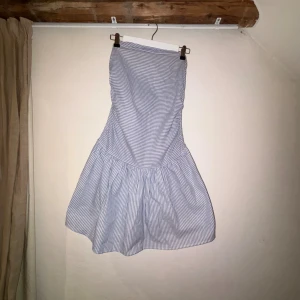 Blåvit randig axelbandslös klänning - Supersöt kort klänning med blåvita ränder och volangig kjol. Axelbandslös modell som sitter snyggt över bysten och har en lätt utställd form. Perfekt för varma dagar och ger en fräsch sommarkänsla. Tunn och skön bomullskänsla i materialet. Säljer då den är förstor för mig över bysten. Dne är köpt på barnavdelningen, men passar XS-S.