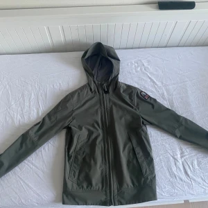 Olivgrön softshelljacka med huva från Parajumpers - Säljer en olivgrön softshelljacka från Parajumpers med huva och dragkedja. Jackan har två snedställda fickor framtill och detaljer i orange på insidan. Perfekt för dig som vill ha en stilren och funktionell jacka.