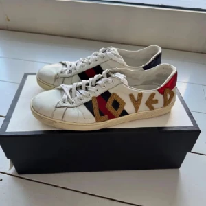 Gucci Loved sneakers i skinn - Gucci sneakers i vitt skinn med guldtexten 'LOVED' på sidan, klassiska röda och marinblå ränder samt metallicröd och marinblå häl. Skorna har snörning och rund tå. Kommer med originalbox och dustbag.