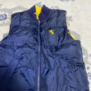 Mörkblå dunväst med gul detalj - Snygg mörkblå dunväst med gul lejonlogga på bröstet och gula detaljer på insidan av kragen. Västen har dragkedja framtill och två snedställda fickor. Perfekt för lager-på-lager och sportig stil.