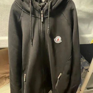 Svart hoodie från Moncler med dragkedja - Svart hoodie från Moncler med hel dragkedja framtill, snörning i huvan och broderad logga på bröstet. Tröjan har två fickor med dragkedja och en broderad detalj på huvan. Perfekt för en chill och stilren look.