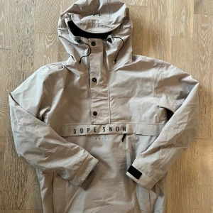 Beige vindjacka från Dope Snow S - Snygg beige vindjacka från Dope Snow med huva och svart snörning. Jackan har tryckknappar framtill, dragkedja i sidan och justerbara muddar med kardborre. Stort Dope Snow-tryck över bröstet. Perfekt för dig som gillar streetwear och outdoor vibes.