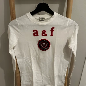 Vit långärmad tröja Abercrombie kids *M* - Vit långärmad tröja från Abercrombie med röd a&f-logga och broderad patch på bröstet. Tröjan har rund halsringning och är gjord i mjuk bomull. Perfekt för en chill och stilren look.