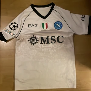 Napoli tröja storlek s - Vit Napoli tröja med snyggt mönster. Tröjan är storlek s och är sparsamt använd. Tröjan har champions league badge på armen. Tveka inte på att höra av er vid funderingar