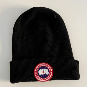 Svart mössa från Canada Goose - Svart ribbstickad mössa från Canada Goose med klassisk uppvikt kant och det ikoniska runda Arctic Program-märket framtill i rött, vitt och blått. Inköpt på Johnells - kvitto finns på mail vid förfrågan. 
