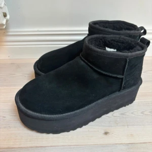 Svarta UGG boots med platåsula - Svarta boots från UGG med mjuk fårskinnsinsida och kraftig platåsula. Skorna är i mocka och har rund tå samt dragflik baktill. Perfekta för kalla dagar när du vill ha både värme och stil. (FRÅGA GÄRNA OM FLER BILDER!!)