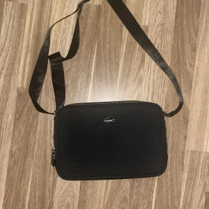 Svart axelväska från Lacoste - Snygg svart axelväska från Lacoste med präglad logga på framsidan och silverfärgad dragkedja. Väskan har justerbar axelrem och är tillverkad i slitstark syntet. Perfekt storlek för det viktigaste och stilren design med diskret krokodillogo.