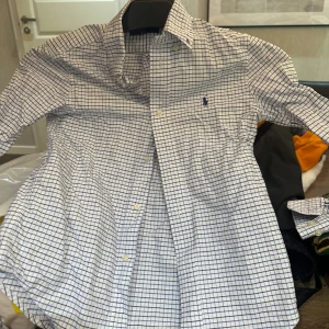 Rutig skjorta från Ralph Lauren (barn, 130) - Snygg vit skjorta med blå rutigt mönster från Ralph Lauren. Klassisk krage, långärmad och med knappar framtill. Ikoniska Polo-loggan broderad på bröstet. Skjortan är gjord i mjuk bomull och har en clean, normal passform som funkar till många stilar.