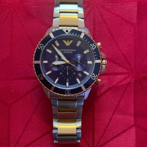 Emporio Armani klocka blå/guld/silver - Snygg klocka från Emporio Armani med blå urtavla, gulddetaljer och silverfärgad länk i metall. Klockan har kronograf, datumvisning och tydliga indexmarkeringar. Boetten är rund med guldfärgad kant och robust känsla. Perfekt accessoar för dig som gillar stilren design. NY PRIS 4600kr även boxen medföljer och länkarna som jag tagit av för den ska passa mig 