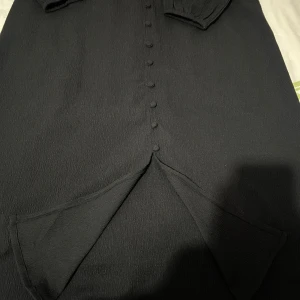 Svart långklänning med knappar - Stilren svart långklänning med långa ärmar och klädda knappar längs hela framsidan. Klänningen har en slits framtill och är tillverkad i ett lätt strukturerat tyg som ger en snygg och modern känsla. Perfekt för dig som gillar minimalistisk och elegant stil.