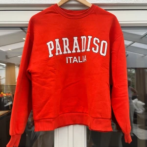 Röd sweatshirt Paradiso Italia - Cool röd sweatshirt med trycket 'PARADISO ITALIA' i vitt framtill. Tröjan har rund halsringning, ribbade muddar och lång ärm. Perfekt för dig som gillar en sportig och avslappnad stil.