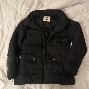 Svart pufferjacka från Cubus - Svart pufferjacka från Cubus i storlek 128. Jackan har dragkedja framtill, två fickor med tryckknappar och hög krage. Perfekt för kyliga dagar och enkel att matcha med allt. Fodrad och riktigt skön att ha på sig.