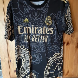 Real Madrid x Adidas svart draktröja M - Säljer en svart Real Madrid fotbollströja från Adidas med guldiga detaljer och stor drakdesign över hela tröjan. Tröjan har korta ärmar, rund hals och är tillverkad i lätt funktionsmaterial. Stora loggor och 'Emirates Fly Better' tryck på bröstet.