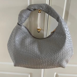 Grå axelväska med knut och reptilmönster nyskick  - Snygg grå axelväska med coolt reptilmönster och en unik knut på handtaget. Väskan har en mjuk, rundad form och stängs med dragkedja i guld. Perfekt storlek för att få plats med allt du behöver. Materialet är syntet och ger en lyxig känsla.