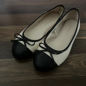Svartvita ballerinaskor från Primark - Stilrena ballerinaskor från Primark i svart och vitt med rund tå och dekorativ rosett framtill. Skorna har svart tåparti och kant, medan resten är i ljus textil. Perfekta för dig som gillar klassisk och enkel stil.