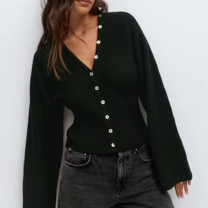 Svart kofta från Gina Tricot - Svart kofta med v-ringning från Gina Tricot. Modellen är croppad med bred ribbad midja och långa ärmar. Snygga silvriga knappar framtill ger en cool detalj. Perfekt till jeans eller kjol för en trendig look. Helt ny! Prislappen kvar