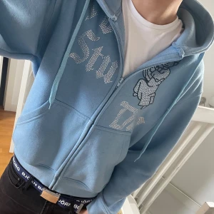 Blå hoodie Von studio med strass - Säljer en ljusblå hoodie från Von studio med dragkedja, huva och snörning. Framsidan har coola strasstenar i text och en ängel. Mjuk bomullskänsla och ribbade muddar. Perfekt för dig som vill sticka ut. Har lite små fläckar men inte så synliga