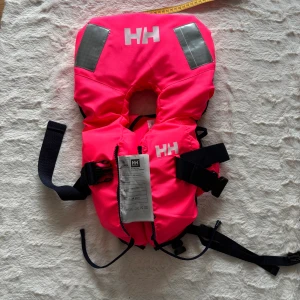 Rosa flytväst från Helly Hansen - Säljer en knallrosa flytväst från Helly Hansen med reflexdetaljer och svarta justerbara remmar. Västen har en hög krage för extra flytkraft och säkerhet, samt spännen och visselpipa. Perfekt för vattenaktiviteter och båtturer. Helt oanvänd, perfekt skick