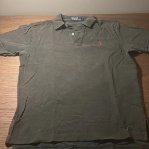 Grå pikétröja från Ralph Lauren - Klassisk grå pikétröja från Polo Ralph Lauren i bomull. Tröjan har korta ärmar, krage med två knappar och den ikoniska röda broderade loggan på bröstet. Perfekt för en stilren och avslappnad look.