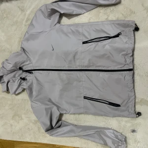 Ljusgrå vindjacka från Nike - Säljer en stilren ljusgrå vindjacka från Nike med svarta dragkedjor och justerbar huva. Jackan har två snedställda fickor med dragkedja och är perfekt för blåsiga dagar. Materialet är lätt och känns sportigt, med klassisk Nike-logga på bröstet.
