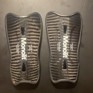 Monolit benskydd i kolfiber - Säljer ett par svarta Monolit benskydd i kolfiber med ergonomisk form och vadderad insida för extra komfort. Lätta och smidiga med snyggt kolfibermönster och vit logga. Perfekta för dig som vill ha maxad prestanda på planen.