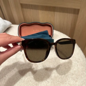 Svarta Gucci solglasögon med gröna detaljer - Knappt använda Snygga svarta solglasögon från Gucci med fyrkantig form och gröna skalmar med röd rand och guldfärgad GG-logga på sidan. Kommer med originalfodral och putsduk. Perfekt accessoar för dig som vill ha en lyxig och trendig look. Köpt för 2500