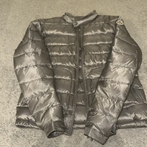 Moncler acorus jacka  - Snygg grå dunjacka från Moncler med glansig finish och horisontella sömmar. Jackan har hög krage med knapp, två fickor med dragkedja och klassisk Moncler-logga på ärmen. Perfekt för kyliga dagar och ger en clean, modern look.