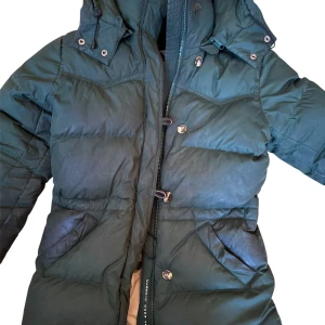 Mörkgrön dunjacka från Boomerang XS - Snygg mörkgrön dunjacka från Boomerang med huva, dragkedja och knappar framtill. Jackan har två stora fickor och även två innerfickor och justerbar dragsko vid huvan. Fylld med äkta dun för extra värme och har en klassisk, puffig look. Perfekt till vintern. Nypris:5999kr