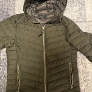 Olivgrön dunjacka från Peak Performance - Snygg olivgrön dunjacka från Peak Performance med huva och dragkedja framtill. Jackan har quiltad design, två fickor med dragkedja och är lätt vadderad för extra värme. Perfekt för kyliga dagar och har en clean, sportig look.