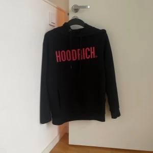 Svart HOODRICH hoodie med röd logga - Svart hoodie från HOODRICH med stor röd logga framtill. Klassisk huva med snörning och magficka. Långärmad och perfekt för en chill streetwear-look. Skön och stilren, passar till allt.