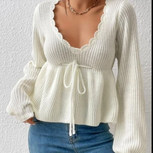 Vit stickad tröja med volangkant - Superfin vit stickad tröja med djup v-ringning och volangkant längs halsen. Tröjan har långa ärmar, knytband i midjan och en lätt utsvängd passform nedtill. Perfekt för dig som gillar en söt och trendig look.