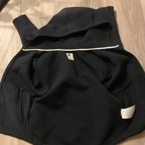 Svart fleecejacka med huva från Nike - Svart fleecejacka från Nike med dragkedja framtill och två sidofickor. Jackan har en stor huva och en vit detalj längs insidan av axlarna. Perfekt för kyliga dagar och enkel att matcha med det mesta.