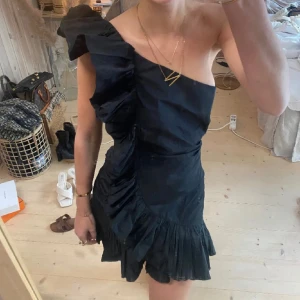 Svart one shoulder volangklänning - Supersnygg svart kort klänning med one shoulder och stora volangdetaljer längs axeln och nederkanten. Klänningen har en asymmetrisk design och är tillverkad i ett lätt, glansigt material som ger en cool och trendig look.