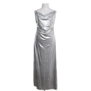 Silverfärgad långklänning med drapering - Snygg långklänning i metallic silver med draperad urringning och smala axelband. Klänningen har en slät, glansig yta och en elegant, figurnära passform som ger en futuristisk vibe. Perfekt för dig som vill sticka ut med en cool och trendig look.