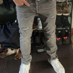 Grå  jeans med slitningar - Säljer ett par grå  jeans från Jack & Jones med snygga slitningar och trasiga detaljer på benen. Jeansen har fem fickor, normal midja och är tillverkade i stretchig bomullsdenim för en tight passform. Perfekta för dig som gillar en trendig och avslappnad stil.
