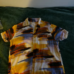 Färgstark kortärmad skjorta från Levi's - Säljer en riktigt cool kortärmad skjorta från Levi's i storlek L. Skjortan har ett färgstarkt abstrakt mönster i gult, blått, orange och svart, med bröstficka och relaxed fit. Perfekt för dig som vill sticka ut med en unik stil.