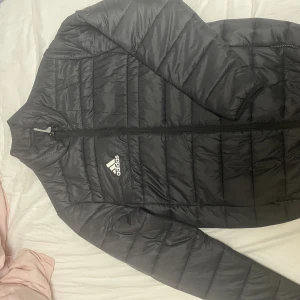 Svart pufferjacka från Adidas S - Svart pufferjacka från Adidas med klassisk logga på bröstet. Jackan har quiltad design, dragkedja framtill och är tillverkad i lätt och smidig polyester. Perfekt för kyliga dagar och enkel att matcha med allt.