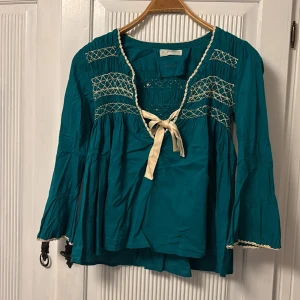 Turkos Odd Molly blus med broderi - Supersöt turkos blus från Odd Molly med broderade detaljer i vitt och dekorativ knytning framtill. Blusen har lång ärm, v-ringning och volangig passform. Perfekt för dig som gillar boho-stil och unika detaljer.