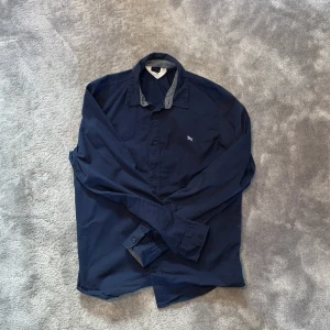 Mörkblå skjorta från H&M - Mörkblå skjorta från H&M med klassisk krage och knäppning framtill. Skjortan har långa ärmar och en liten broderad detalj på bröstet. Perfekt för en stilren och enkel look.