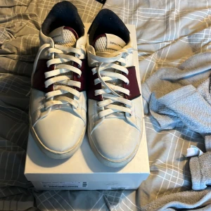 Emporio Armani vita sneakers med röd detalj - Snygga vita sneakers från Emporio Armani med breda röda detaljer över snörningen. Skorna har klassisk rund tå, vita snören och en platt sula. Tillverkade i skinn för en lyxig känsla och passar perfekt till streetwear eller en clean look.